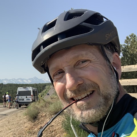 Todd Millard smiling on a gravel road ride, cycling helmet on and sunglasses held in his teeth, with mountains and other riders behind him