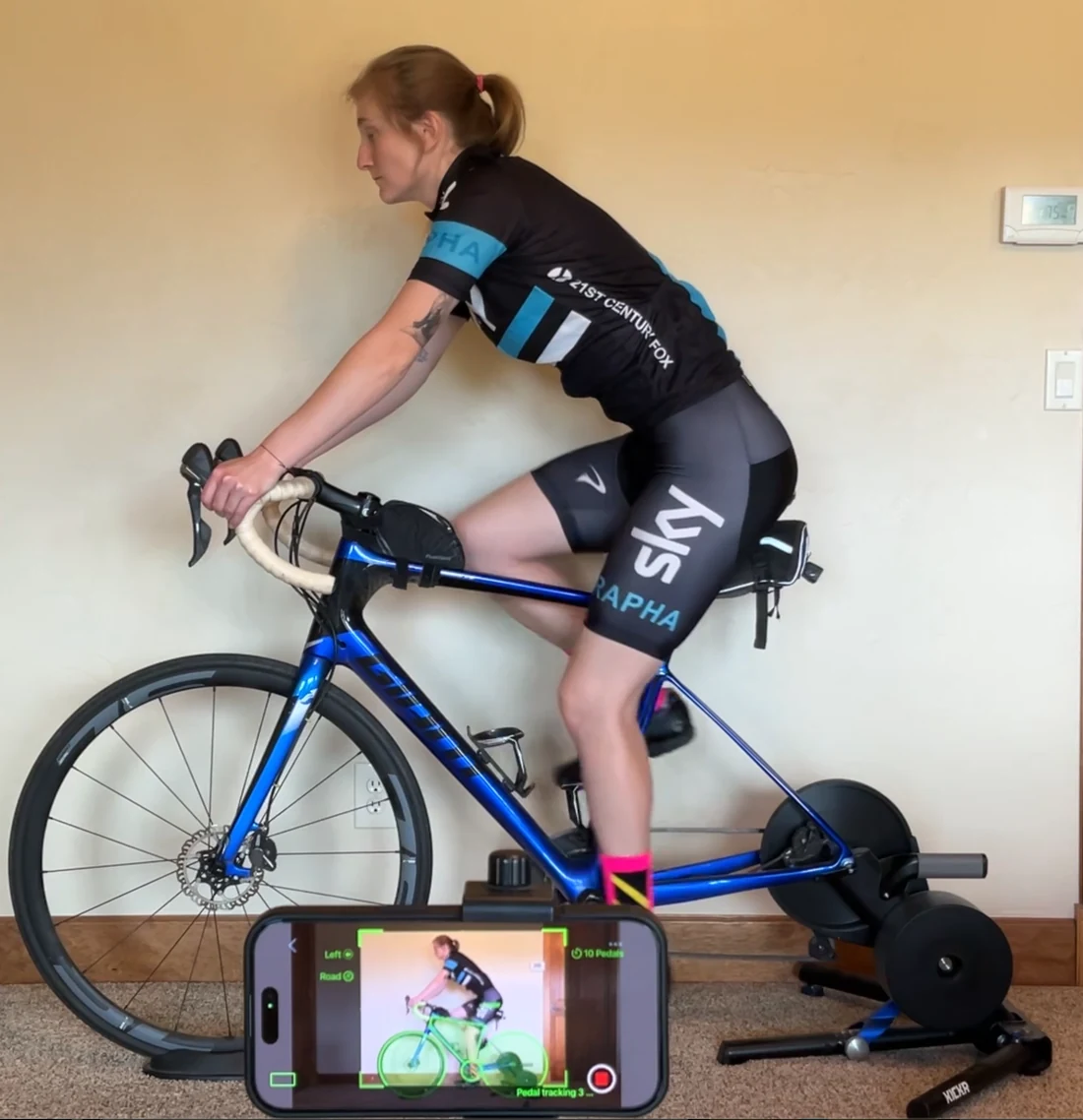 A rider pedaling on an indoor trainer with an iPhone propped in front of her, its screen showing Bike Fast Fit EZ recording with the tracked skeleton locked on and no markers on the rider