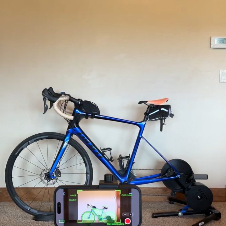 A road bike on an indoor trainer with an iPhone mounted in front of it, the Bike Fast Fit recording screen framing the bike