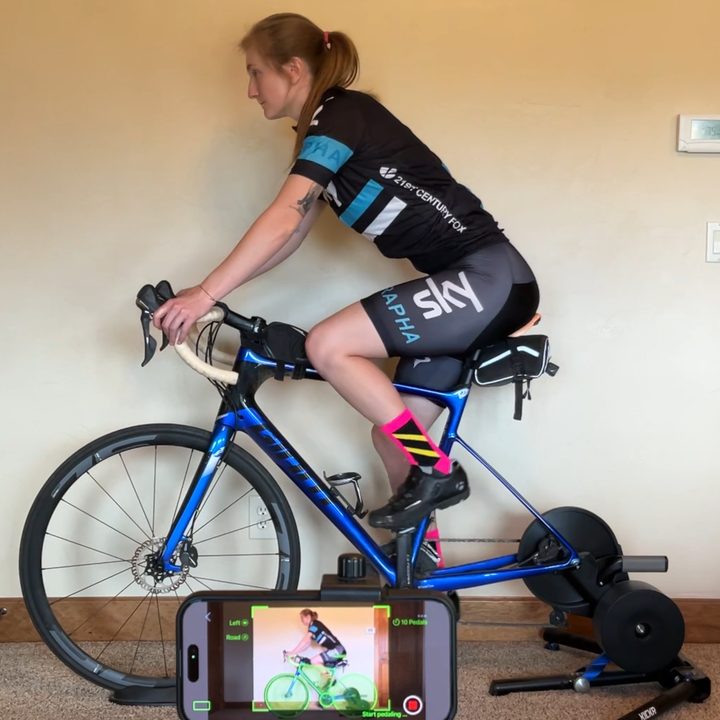 A rider pedaling on the indoor trainer while the mounted iPhone records the side view